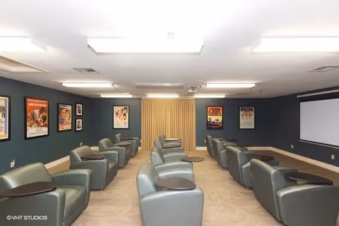 A small theater room with two rows of comfortable gray recliner chairs, each equipped with a small attached table. The walls are dark blue and decorated with framed movie posters. A large projector screen is visible on the right side of the room, and the floor is carpeted in a light beige color. The room is well-lit with ceiling lights.