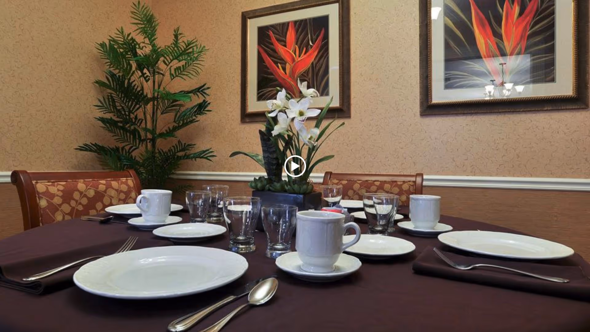 A dining table set with white plates, cups, saucers, glasses, and silverware on a dark tablecloth in a room with patterned wallpaper. There is a floral centerpiece on the table and two framed floral paintings on the wall behind. A green potted plant is visible in the corner.