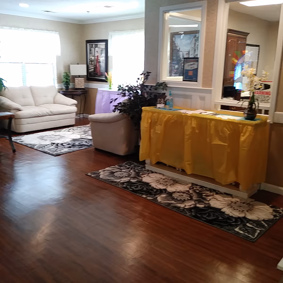 A cozy living room area with wooden flooring, two floral rugs, a white couch, a beige armchair, and a small table covered with a yellow cloth holding hand sanitizer and papers. The room has large windows with white curtains, framed artwork on the walls, and some potted plants.