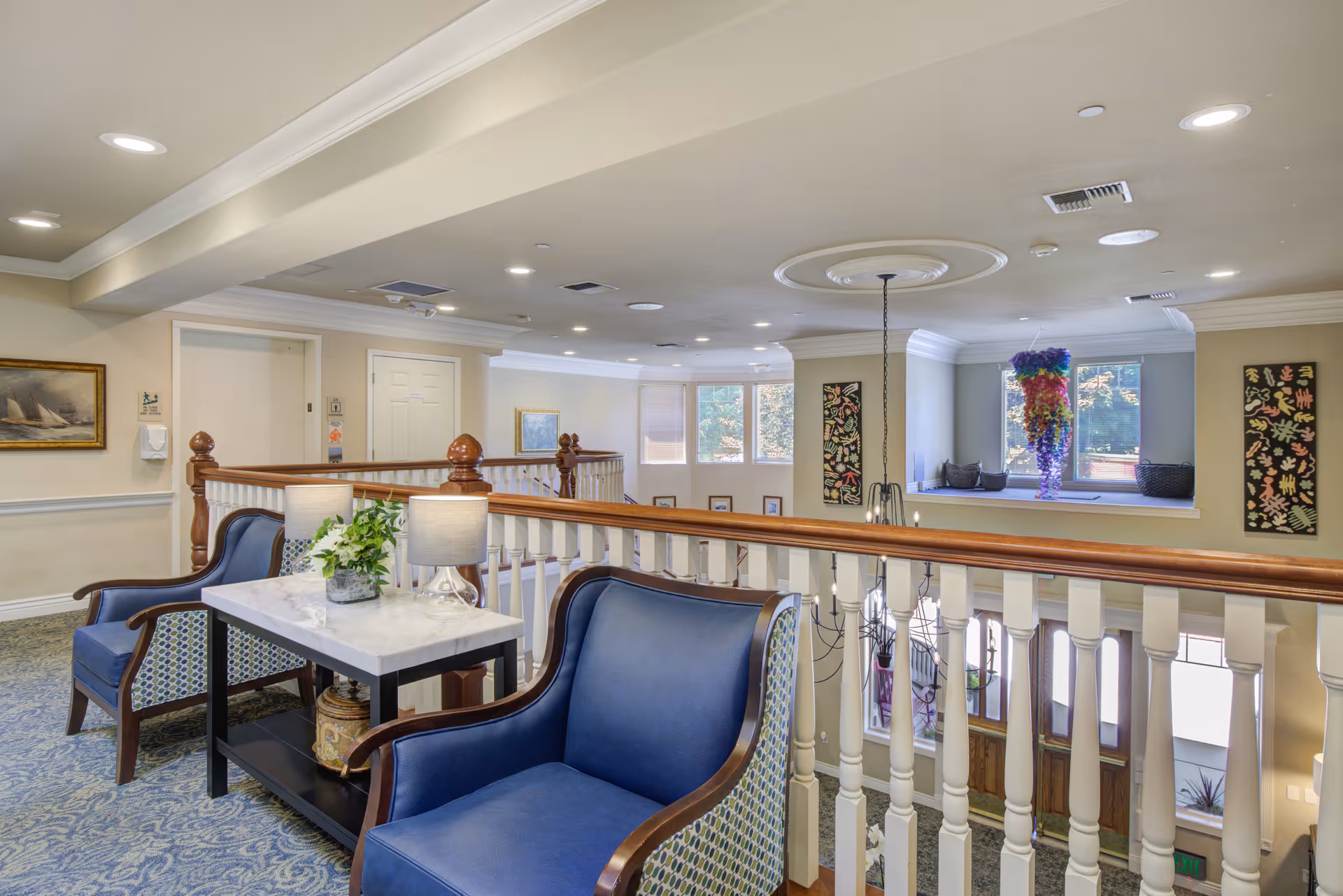 A bright and spacious interior common area with blue upholstered armchairs and a small table with two lamps and a plant. The area overlooks a lower level with large windows and a hanging colorful decoration. The space features carpeted floors, white railings with wooden handrails, and framed artwork on the walls.