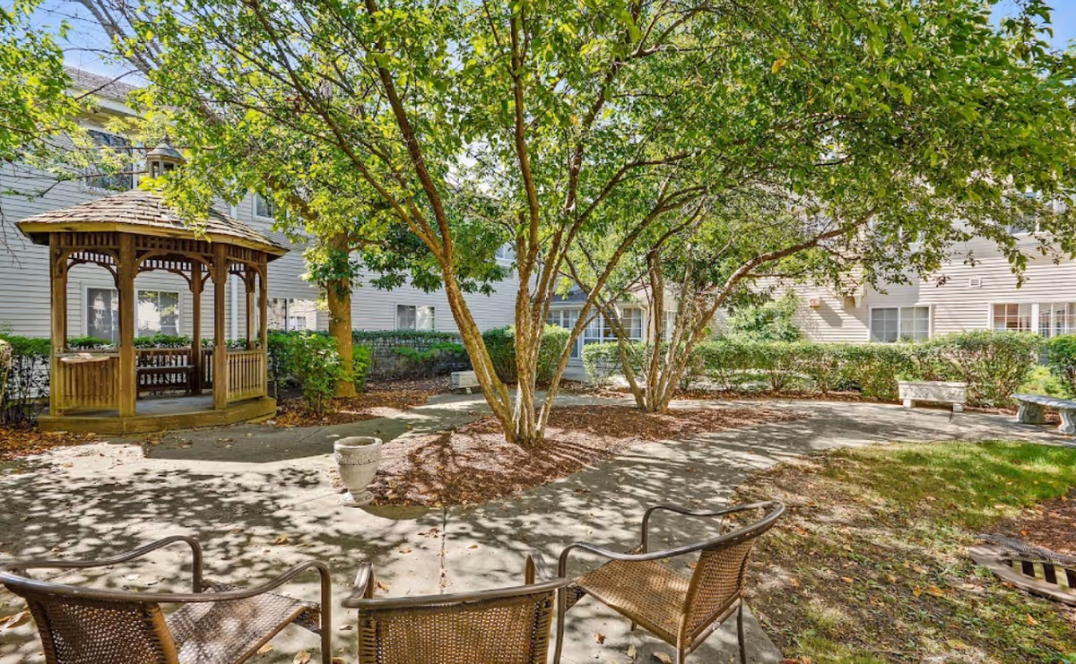 Outdoor courtyard area with a wooden gazebo, several trees providing shade, a concrete pathway, and metal chairs arranged around the space. Surrounding the courtyard are bushes and the exterior walls of a multi-story building with windows.