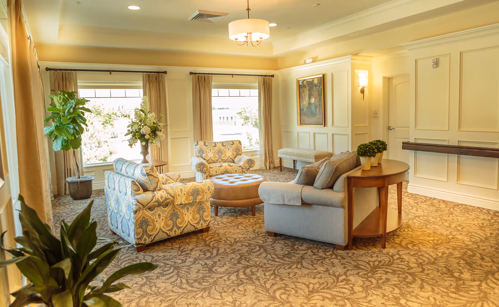 A bright and elegant living room area with patterned armchairs, a gray sofa, a round ottoman, and a wooden side table with small potted plants. The room features large windows with beige curtains, a patterned carpet, wall paneling, a painting, and soft lighting from ceiling and wall fixtures.