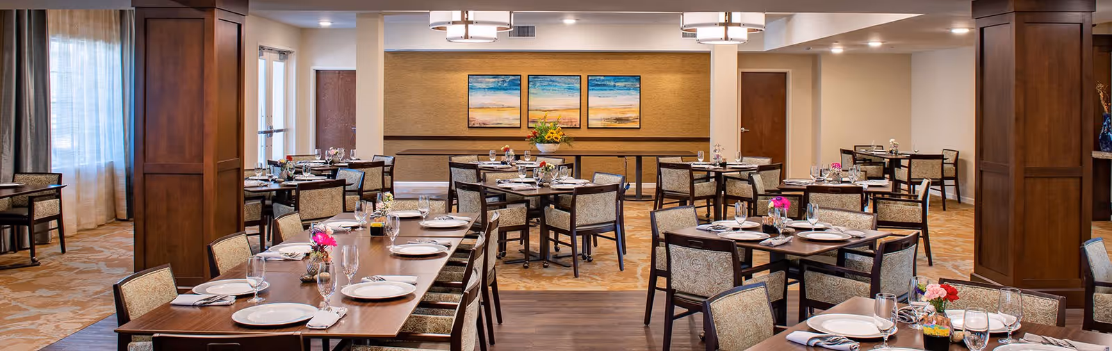 A spacious dining room with multiple tables set for a meal, featuring plates, glasses, napkins, and small floral centerpieces. The room has warm lighting, wooden columns, and three landscape paintings on the far wall. Large windows with curtains allow natural light to enter the space.