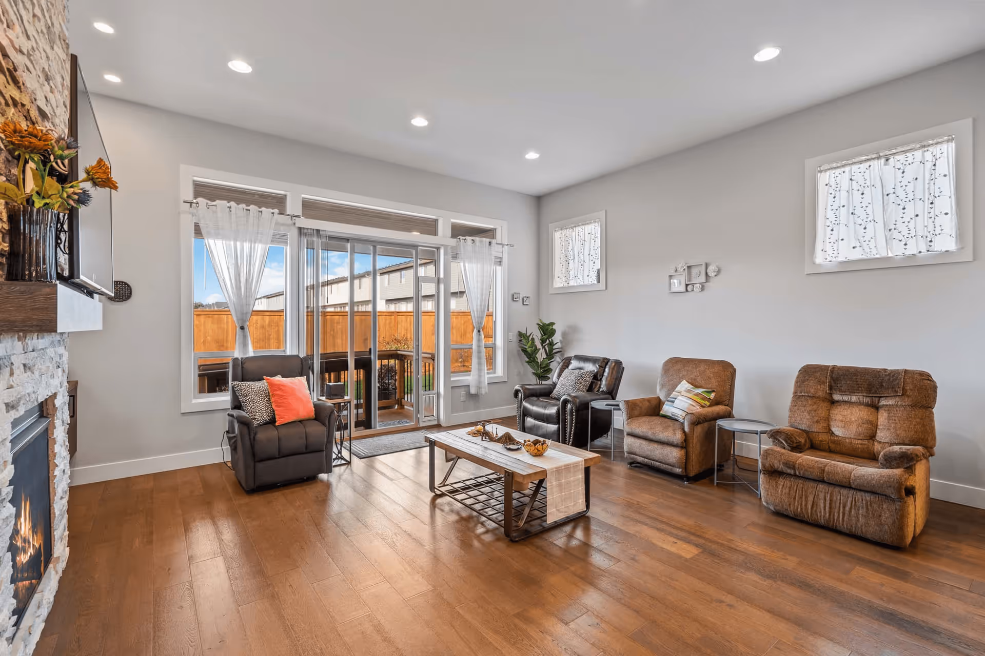 Spacious, well-lit living room with hardwood floors, a stone fireplace, sliding glass doors to the outside, and several recliner chairs arranged around a coffee table.