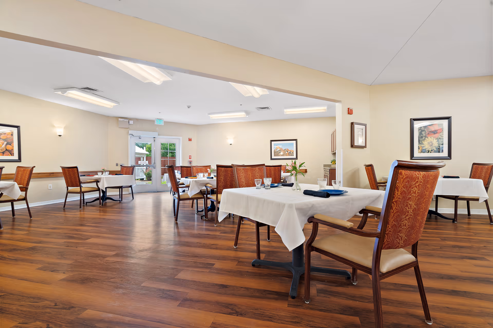 A spacious dining room with wooden floors and several tables covered with white tablecloths. Each table has chairs with patterned upholstery, place settings including plates, glasses, and napkins, and some have small flower vases. The room is well-lit with ceiling lights and wall sconces, and there are framed pictures on the beige walls. A glass door leads outside, allowing natural light into the room.