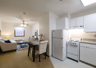 Interior view of a small apartment featuring a compact kitchen with white cabinets, refrigerator, stove, and dishwasher on the right side. Adjacent to the kitchen is a dining area with a small table and two chairs. In the background, there is a living room with a sofa, coffee table, lamp, and a window with a curtain.