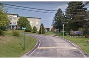 Driveway leading up to a multi-story senior living facility building named St Anne Home, surrounded by trees and greenery under a clear sky.
