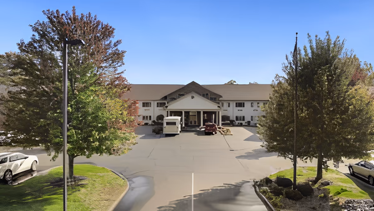 Front exterior view of a large senior living facility building with a covered entrance, surrounded by trees and a parking area with several cars parked.