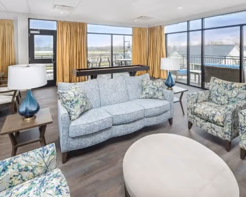 A bright living room with a light blue sofa, two floral patterned armchairs, a round beige ottoman, two side tables with blue glass lamps, large windows with gold curtains, and a door leading to an outdoor balcony with seating.