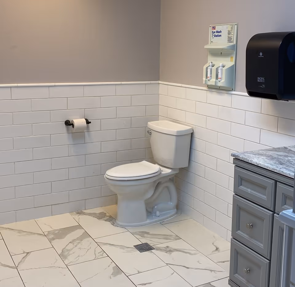 A clean restroom with a toilet against tiled walls, a toilet paper holder, eye-wash station, paper towel dispenser, and a vanity with marble countertop.