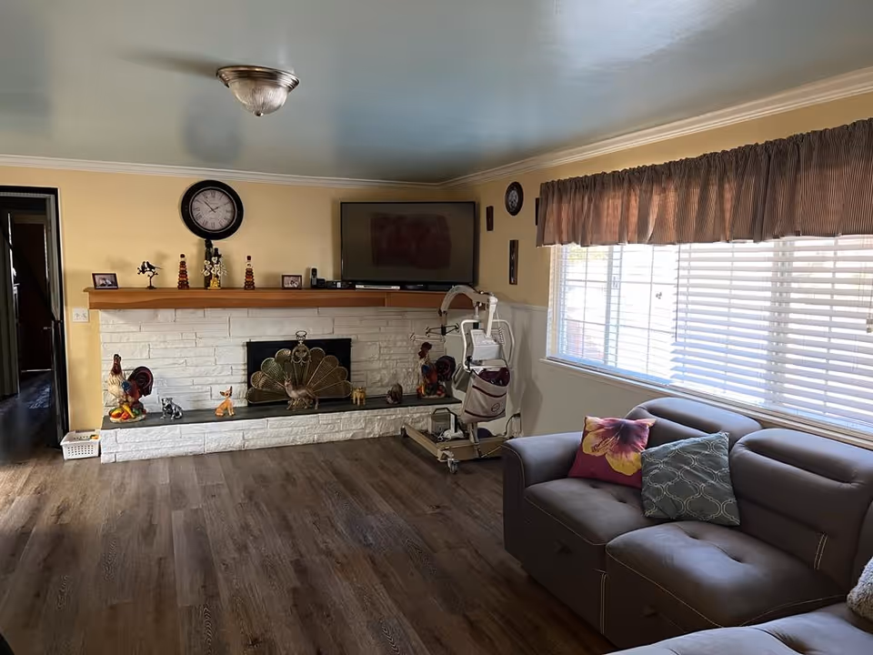 A cozy living room with a gray sectional sofa adorned with colorful pillows next to a large window with blinds and a valance. Across the room is a white brick fireplace with a wooden mantel decorated with small figurines, a clock, and framed photos. Above the mantel is a mounted flat-screen TV. The floor is wood, and there is a ceiling light fixture. A patient lift device is positioned near the fireplace.