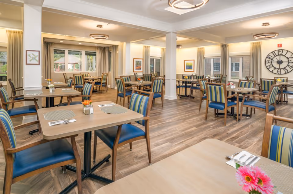 A bright and spacious dining room with multiple wooden tables and chairs featuring blue and green striped cushions. The room has large windows with beige curtains, wooden flooring, and modern circular ceiling lights. Small flower arrangements and place settings are on the tables, and framed artwork and a large decorative clock adorn the walls.