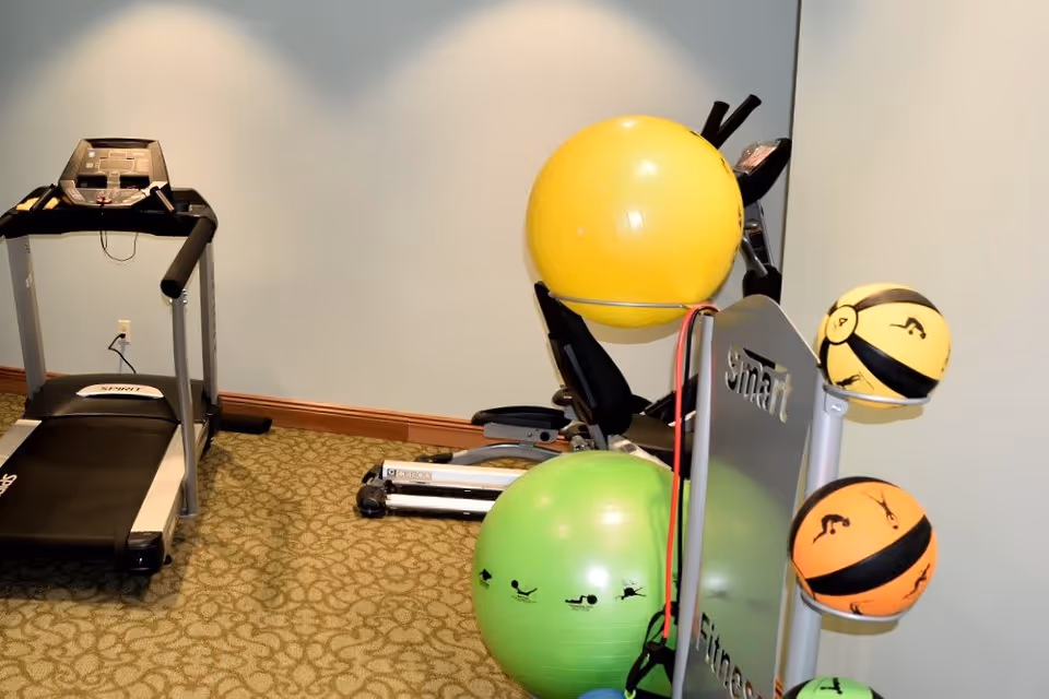 A small fitness room with a treadmill on the left and an exercise bike in the background. In the foreground, there is a rack holding colorful exercise balls and resistance bands. The room has beige patterned carpet and light-colored walls.
