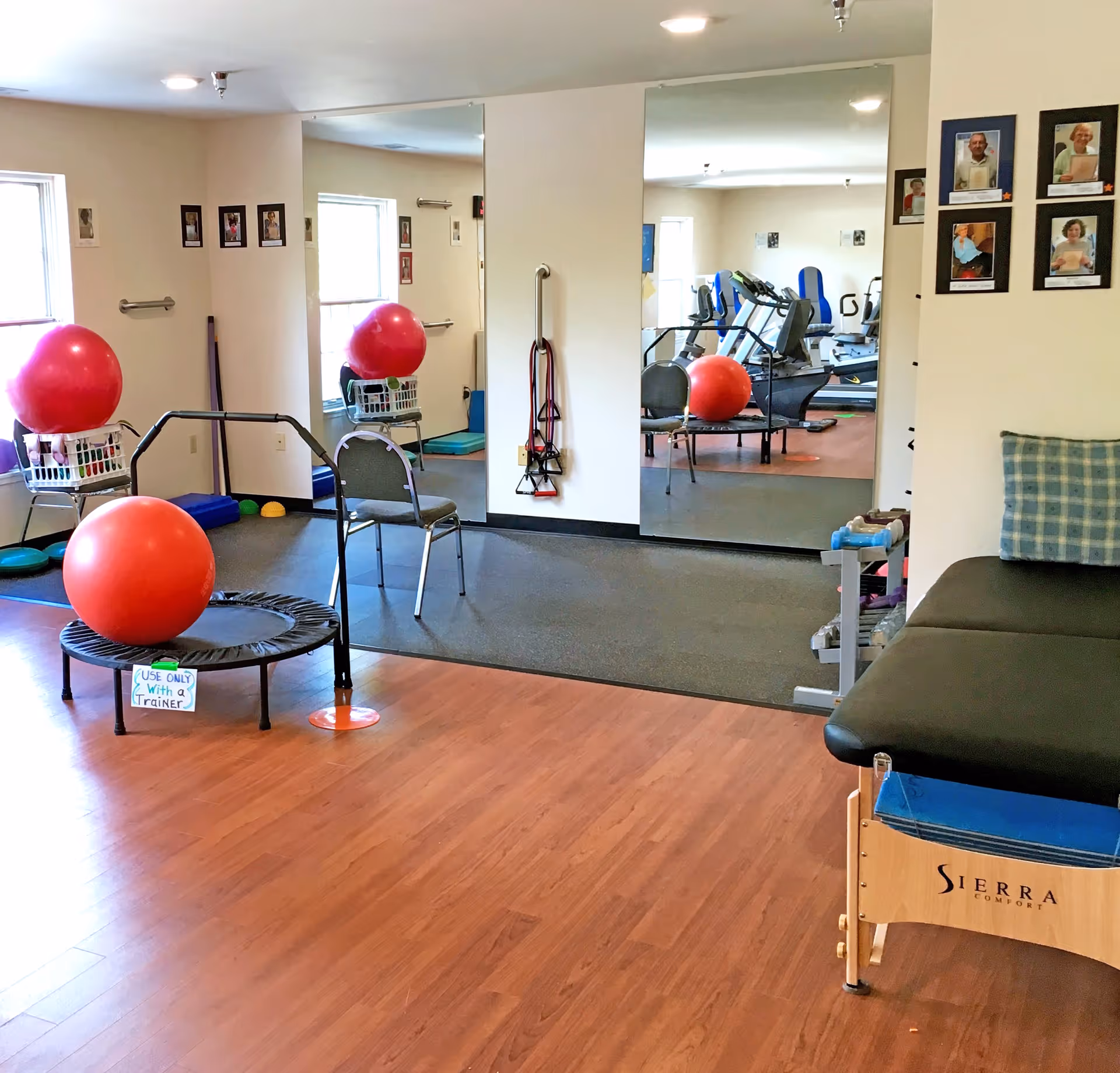 A senior living facility exercise room with wooden flooring and a black rubber mat area. The room contains exercise equipment including a small trampoline with a large red exercise ball on it, a chair, resistance bands hanging on the wall, and a therapy table with a blue cushion. There are large mirrors on the wall reflecting the equipment and room. Several framed photos of people are displayed on the walls.
