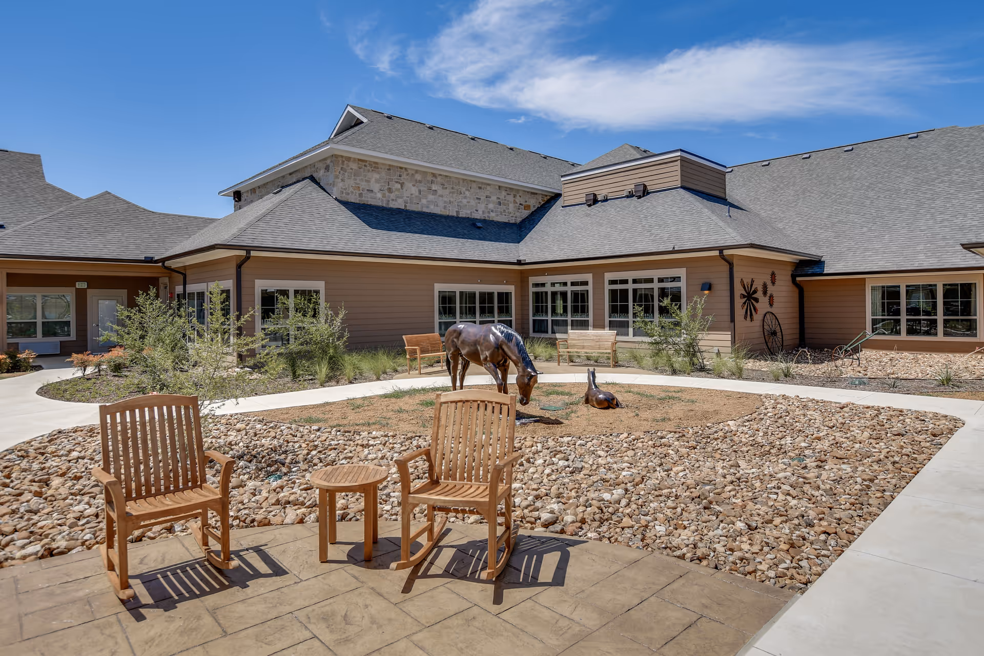 Outdoor courtyard area at Harvest of Roanoke featuring two wooden rocking chairs and a small round table on a paved patio. The courtyard has a dry landscaping design with rocks and sparse plants, and includes sculptures of a horse and a duck. The building surrounding the courtyard has beige siding, multiple windows, and a gray shingled roof under a clear blue sky.