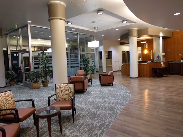 A spacious and well-lit senior living facility lobby area with a mix of carpeted and wood flooring. There are several cushioned chairs with patterned upholstery arranged around small glass-top tables. Large white columns and potted plants add to the decor. In the background, there is a reception desk with wood paneling and a glass partitioned office area.