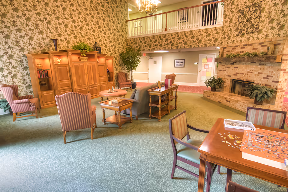A cozy living room area in a senior living facility with floral wallpaper, a brick fireplace, wooden furniture including armchairs, a sofa, side tables, and a table with a partially completed jigsaw puzzle. There is a balcony railing visible on the upper level and plants placed around the room.