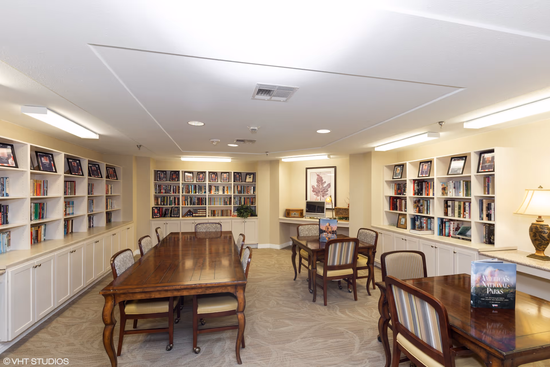 A well-lit library or reading room with multiple wooden tables and chairs arranged for group seating. The walls are lined with white built-in bookshelves filled with books and framed pictures. There is a framed botanical print on the far wall, a table lamp on the right side, and a small TV or monitor on a cabinet at the back. The carpet has a subtle pattern, and the ceiling has recessed lighting.