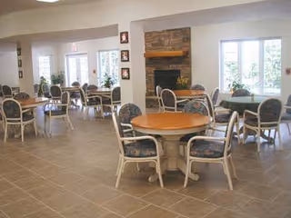 A spacious dining room with several round wooden tables and cushioned chairs arranged around them. The room features large windows allowing natural light to fill the space, a stone fireplace with a wooden mantel, and light-colored tiled flooring.