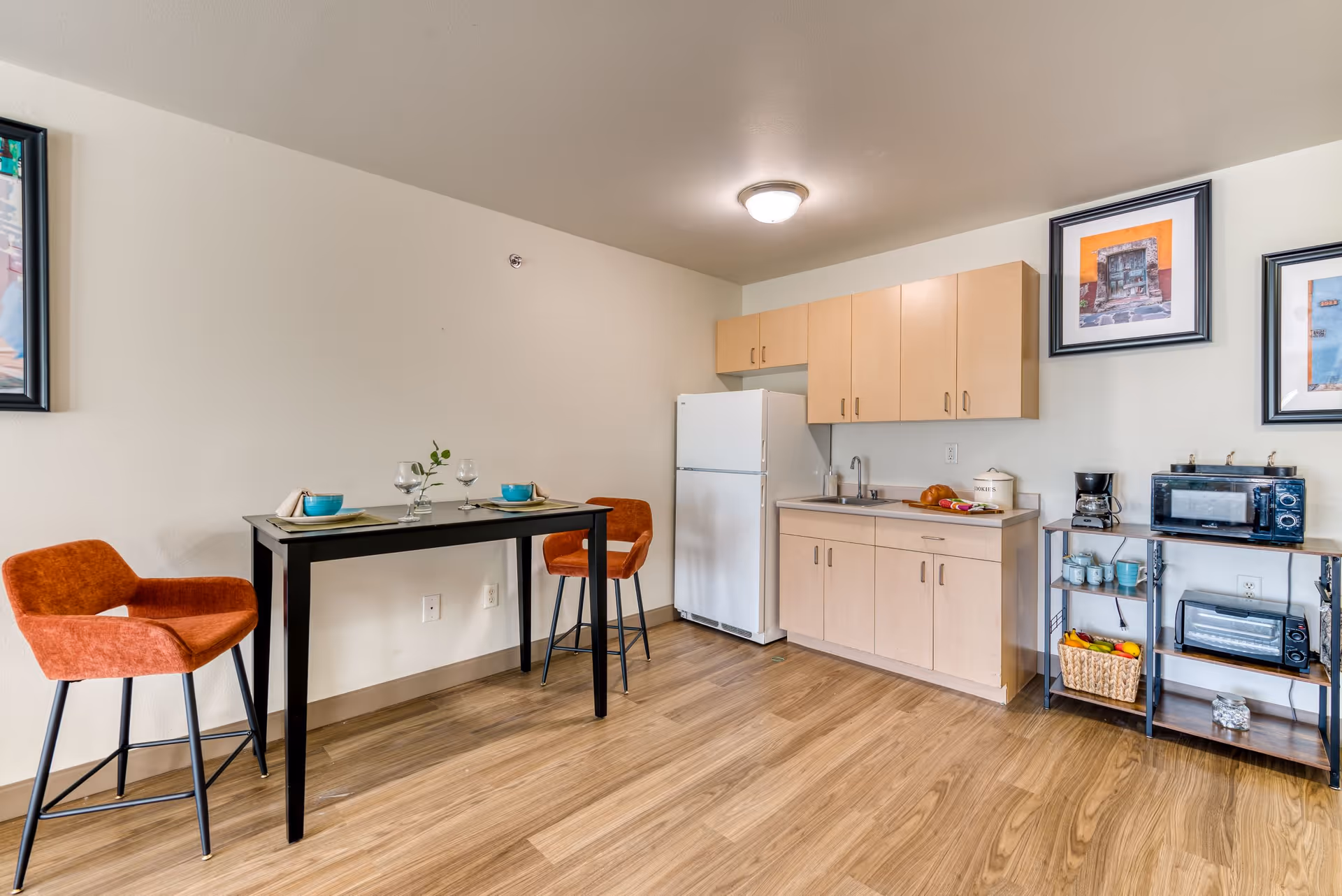 A small kitchen area with light wood cabinets, a white refrigerator, and a countertop with a sink. To the right, there is a metal shelving unit holding a microwave, toaster oven, coffee maker, mugs, and a basket of fruit. On the left side, there is a black dining table set for two with two orange upholstered chairs. The floor is wood, and the walls are light-colored with framed artwork hanging.