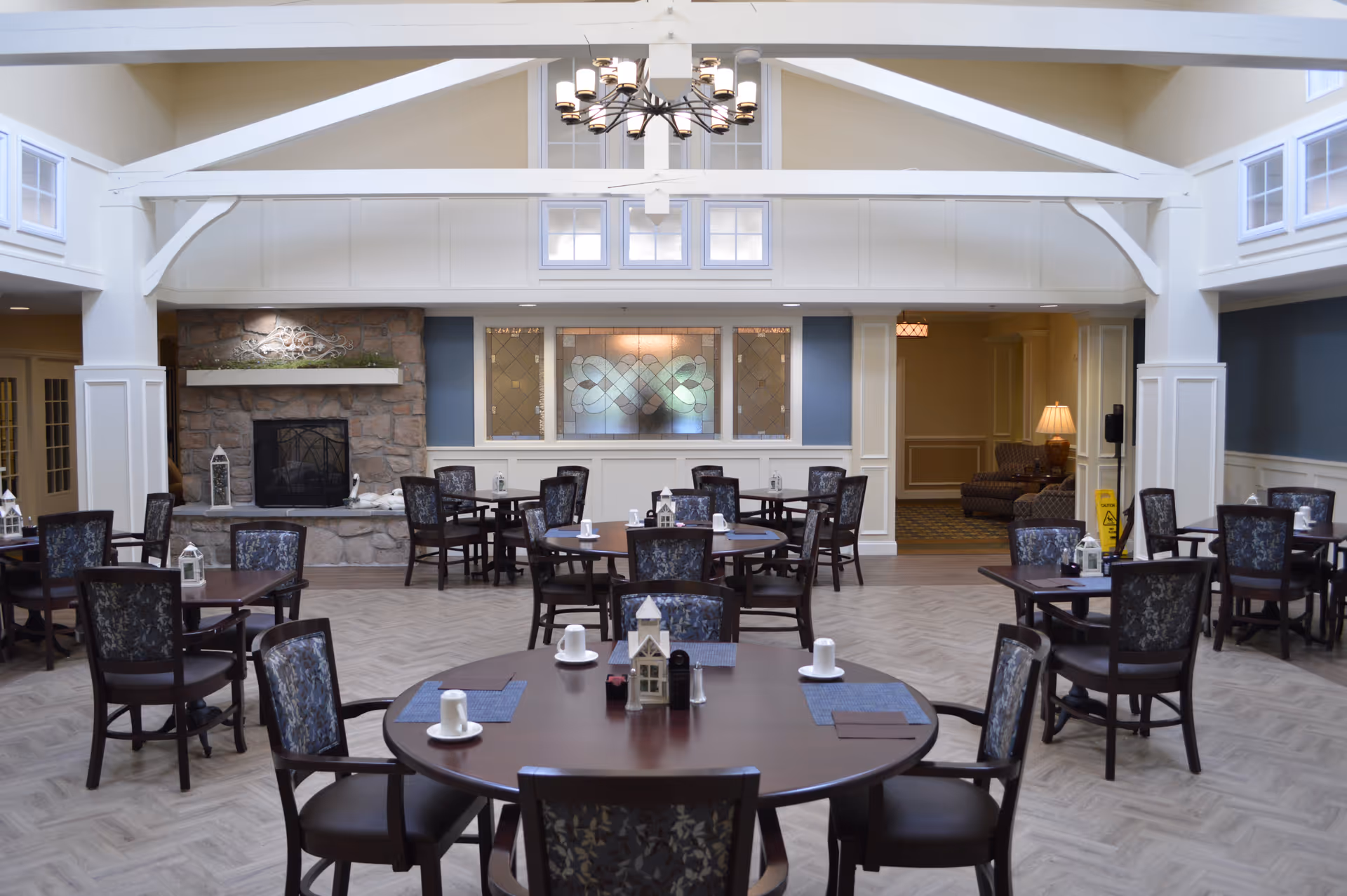A spacious dining room in a senior living facility with multiple round and square tables set with placemats, cups, and decorative lanterns. The room features a stone fireplace on the left wall, a chandelier hanging from a high ceiling with exposed white beams, and a stained glass window in the center background. There is a cozy seating area with armchairs and a lamp visible through an open doorway on the right side.