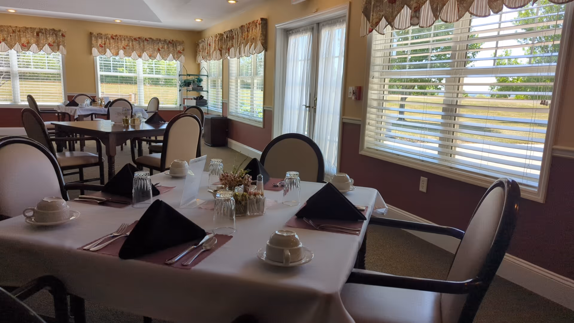 A dining room with several tables set for a meal. Each table has white tablecloths, black folded napkins, cups, glasses, and silverware. The room has large windows with blinds and valances, allowing natural light to fill the space. Chairs with cushioned seats and backs surround the tables.