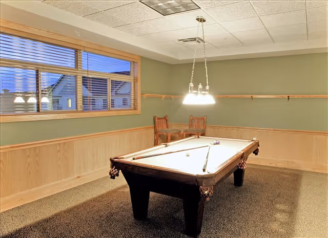A well-lit interior game room featuring a pool table centered under hanging lights, with two chairs and a window in the background.