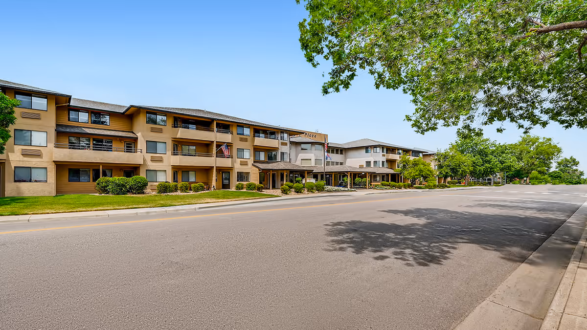 Exterior view of a three-story senior living facility building named Greeley Place with balconies, windows, and a covered entrance. The building is surrounded by green grass, bushes, and trees, with a clear blue sky overhead and a paved road in front.