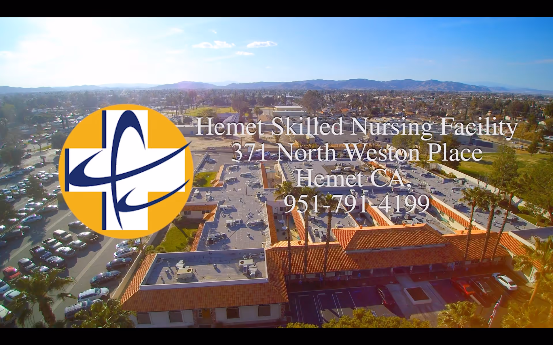 Aerial view of Hemet Skilled Nursing Facility showing multiple buildings with flat roofs and a parking lot filled with cars. The surrounding area includes trees, roads, and distant mountains under a clear sky.