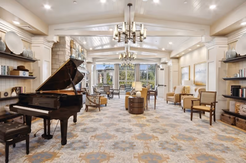 A spacious and elegant common area featuring a black grand piano on the left, several comfortable armchairs and small tables arranged throughout the room, large windows at the far end letting in natural light, decorative shelves with books and vases on the walls, and modern chandeliers hanging from a white paneled ceiling.