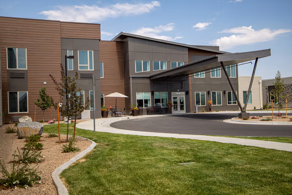 Exterior view of a modern senior living facility building with a covered entrance, large windows, landscaped lawn, small trees, and a clear sky.