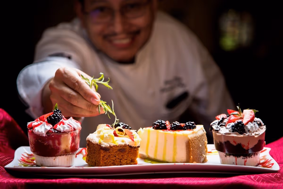 A chef in a white uniform garnishing a plate of assorted desserts, including cakes and parfaits topped with berries and cream, on a red tablecloth.