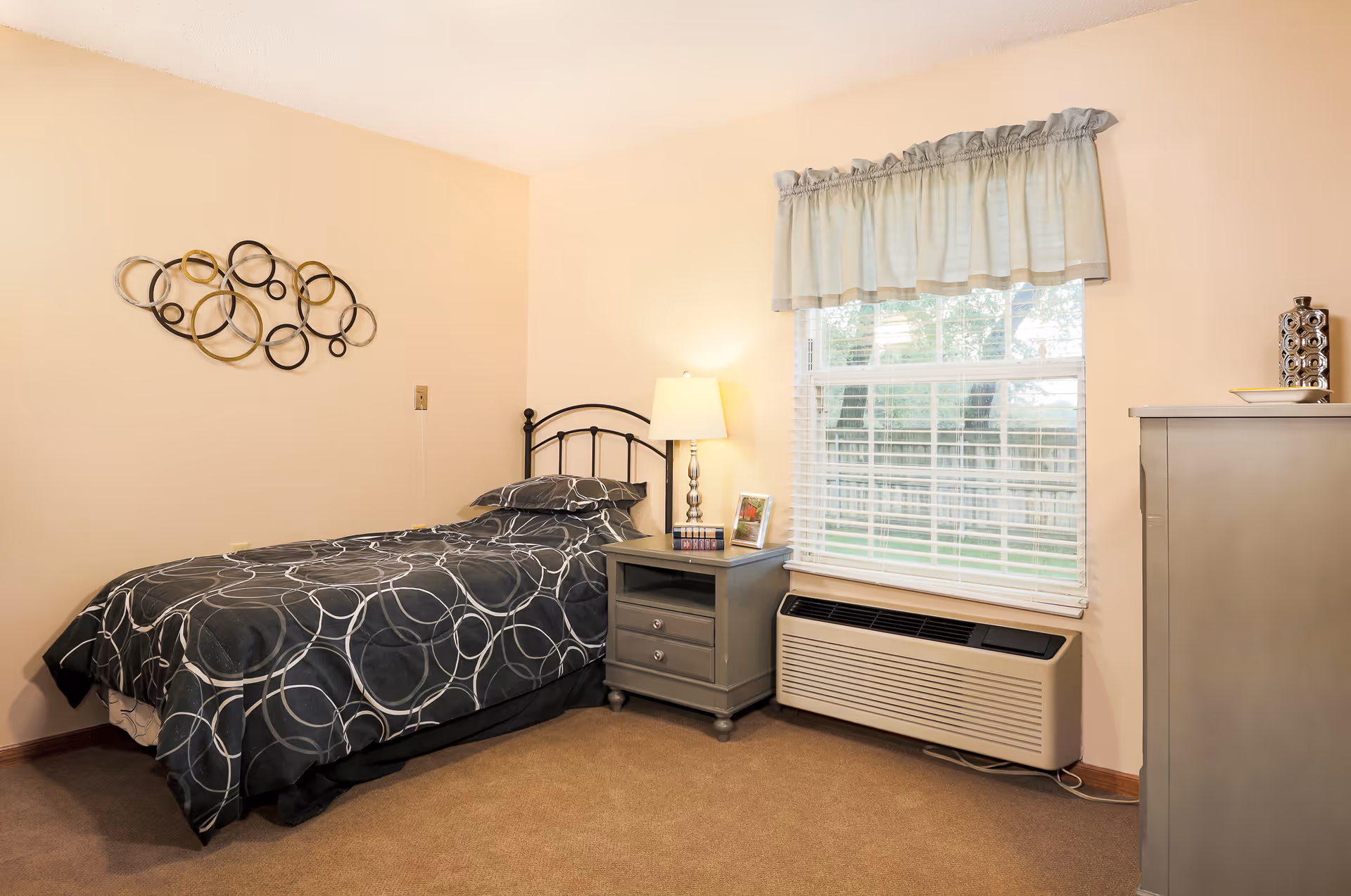 A small bedroom with a single bed covered in a black and white circular patterned bedspread. Next to the bed is a gray nightstand with a lamp, a framed photo, and a small stack of books. A window with white blinds and a light blue valance is on the right wall, above a beige air conditioning unit. A decorative metal wall art piece with circular designs is mounted on the left wall. The room has beige walls and carpeted floor.