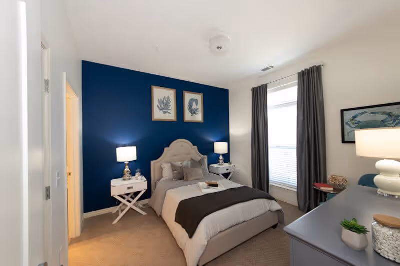 A cozy bedroom with a beige upholstered bed against a dark blue accent wall. The bed is neatly made with gray and beige bedding and several pillows. Two white nightstands with lamps flank the bed. There are two framed botanical prints hanging above the bed. A window with gray curtains is on the right side of the room. A gray dresser with a lamp and small decorative plants is in the foreground on the right side. The room has beige carpeting and white walls.