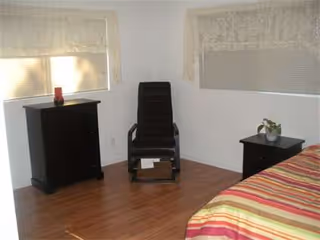 A small bedroom with a striped bed, black dressers, chair, and lace-curtained windows.