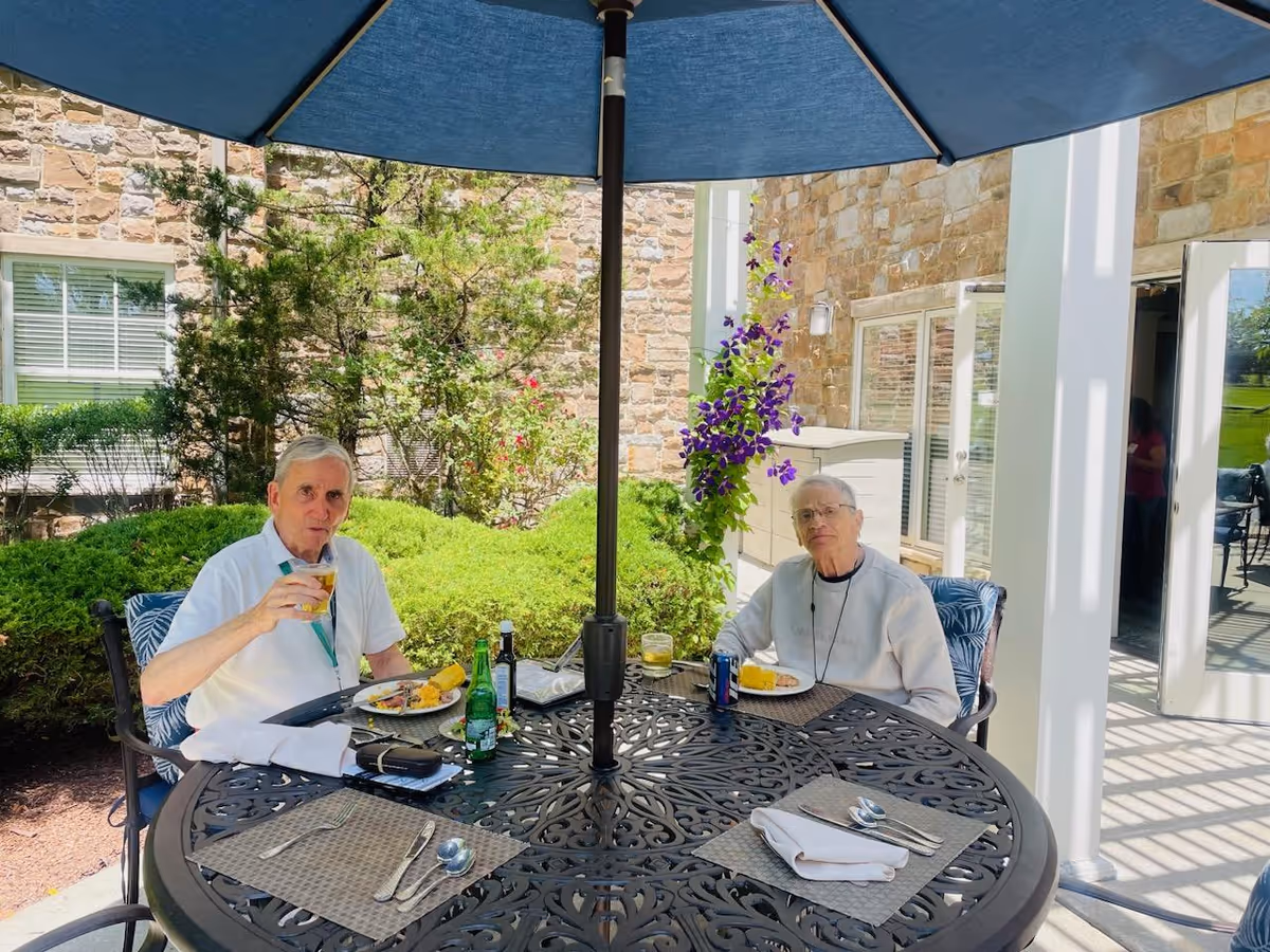 Two elderly men sitting at a round outdoor metal table under a large blue umbrella, enjoying a meal with plates of food and drinks. They are seated in a garden area with green bushes, flowers, and a stone building wall in the background.