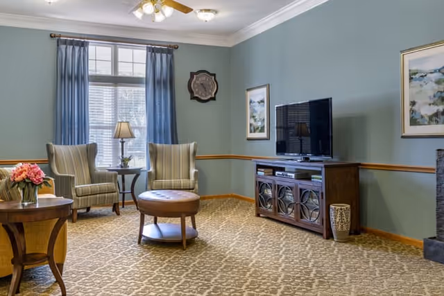 A cozy living room with two striped armchairs and a round ottoman in the center. There is a wooden TV stand with a flat-screen television on it, flanked by a decorative vase. The walls are painted light blue with a wooden chair rail, and there are two framed paintings and a clock on the walls. A window with blue curtains allows natural light to enter the room, and a table lamp sits between the armchairs.