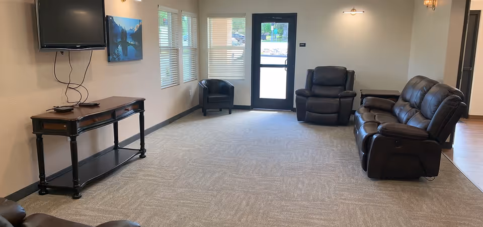 Small lounge with leather recliner chairs and a sofa, wall-mounted TV above a console table, windows, and an exterior door.