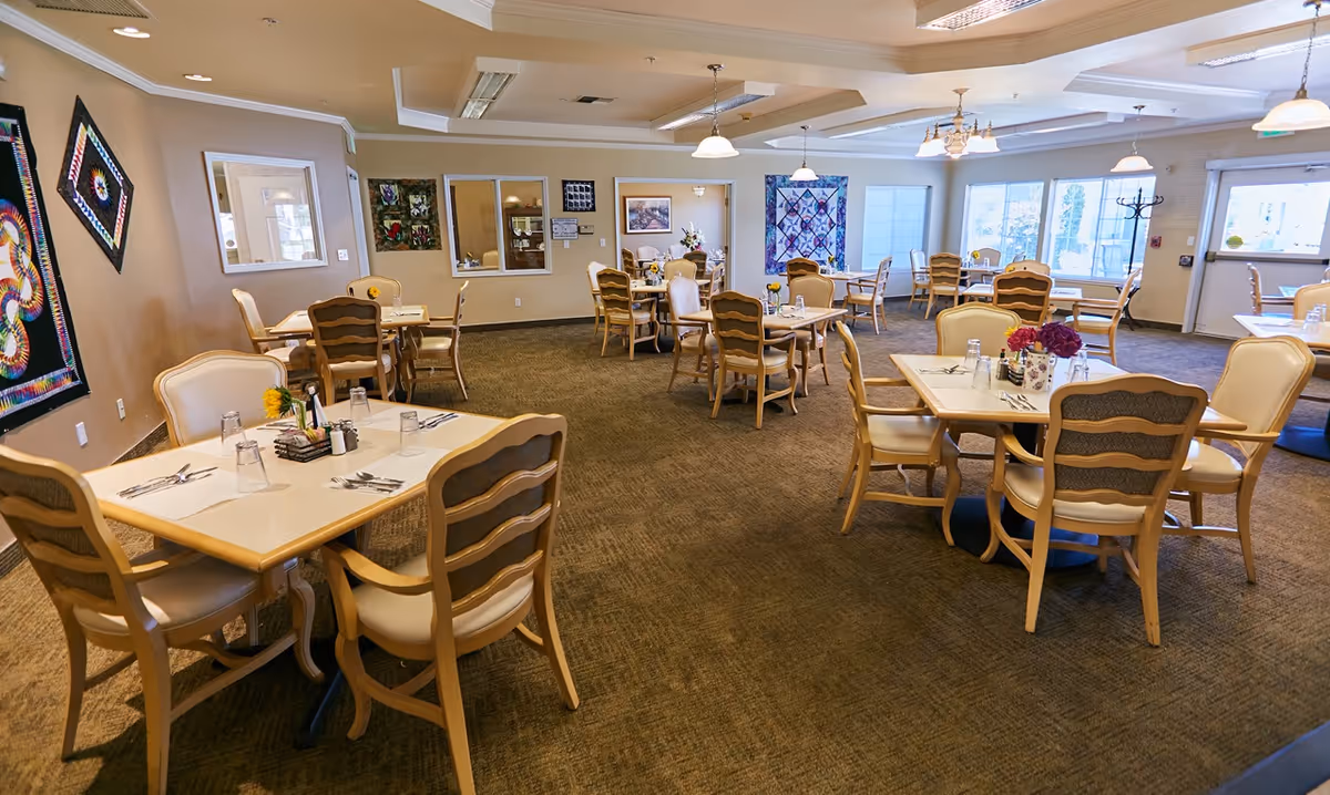 A spacious dining room in a senior living facility with multiple tables and chairs arranged neatly. Each table is set with placemats, utensils, glasses, and small flower vases. The room has beige walls adorned with colorful quilted wall hangings and large windows allowing natural light to fill the space.