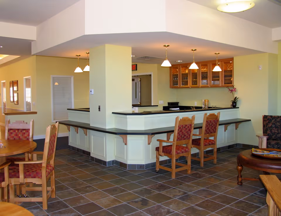 Interior view of a senior living facility common area with a bar counter and several wooden chairs with red patterned cushions. The room has tiled flooring, yellow walls, and hanging pendant lights above the counter. There are glass-front cabinets on the wall behind the counter and a round wooden table with chairs in the foreground.