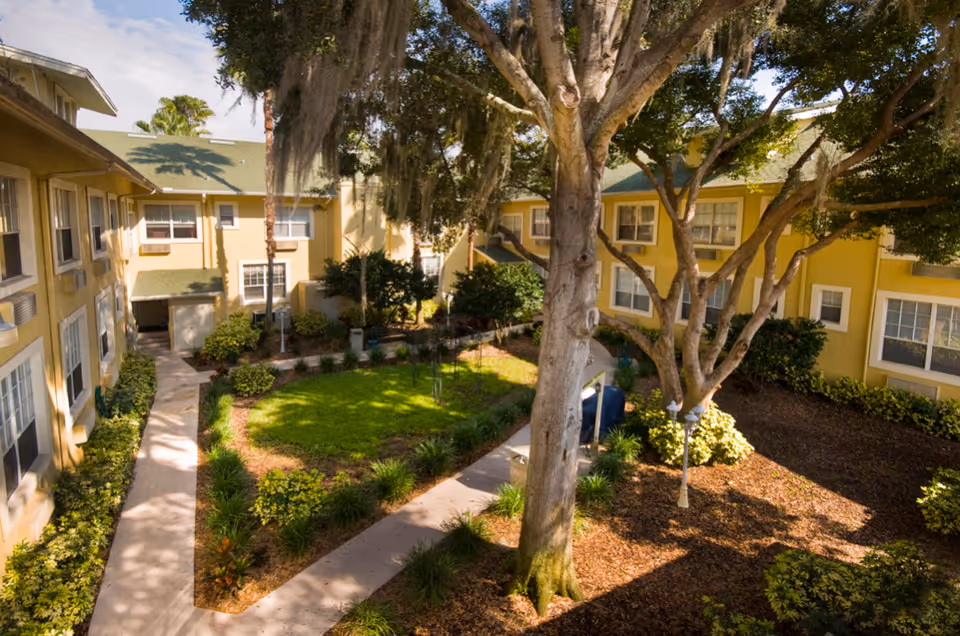 View of a landscaped courtyard area surrounded by a two-story yellow building with green roofs. The courtyard features a large tree, green grass, shrubs, and paved walkways.
