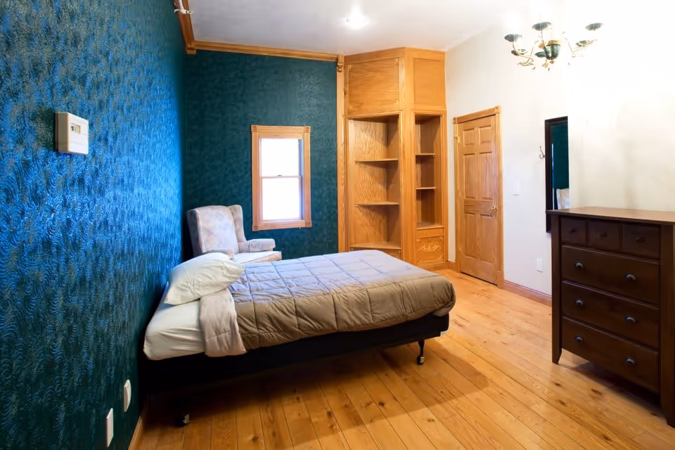 A bedroom with a single bed covered with a beige comforter and white pillows. There is a patterned armchair next to a window with wooden trim. The walls are painted dark green and white, with wooden crown molding. A wooden dresser with drawers and a mirror above it is on the right side, and built-in wooden shelves are in the corner near a closed wooden door. The floor is made of light-colored wood.
