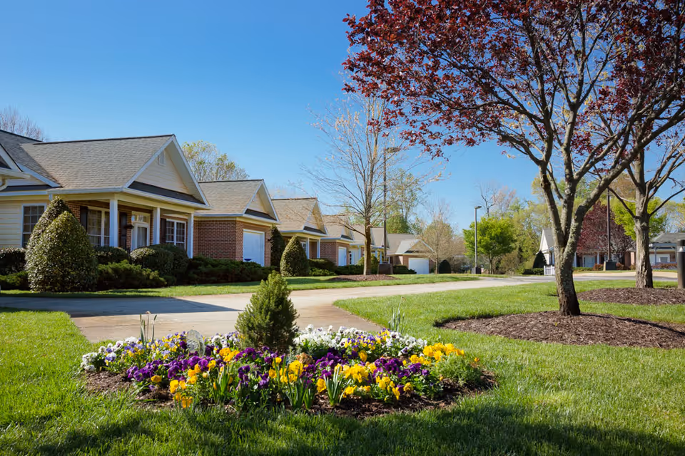 A row of single-story residential buildings with brick and siding exteriors, surrounded by well-maintained green lawns and landscaped flower beds with colorful flowers. Trees with red and green leaves line the sidewalk under a clear blue sky.