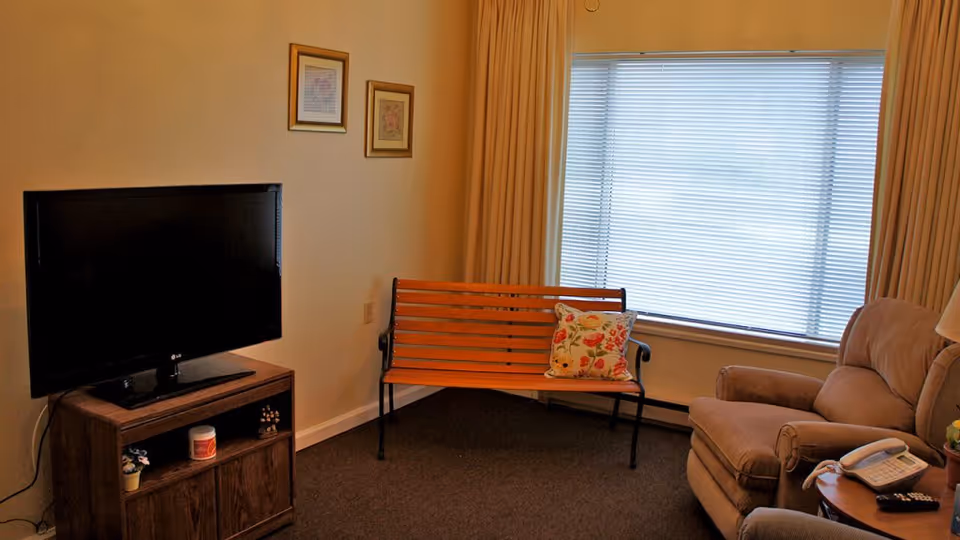A cozy living room area with a flat-screen TV on a wooden stand, a wooden bench with a floral cushion, a large window with closed blinds and beige curtains, and a comfortable armchair next to a small round table holding a telephone and a remote control.