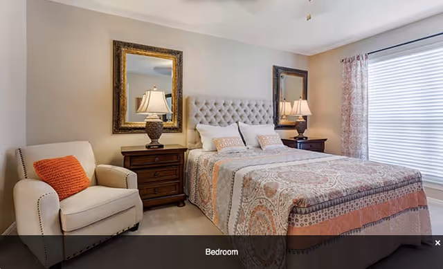 A cozy bedroom featuring a large bed with a patterned bedspread and multiple pillows. On either side of the bed are wooden nightstands with matching lamps and framed mirrors above them. To the left of the bed is a beige armchair with an orange cushion. A window with blinds and patterned curtains is on the right side of the room.