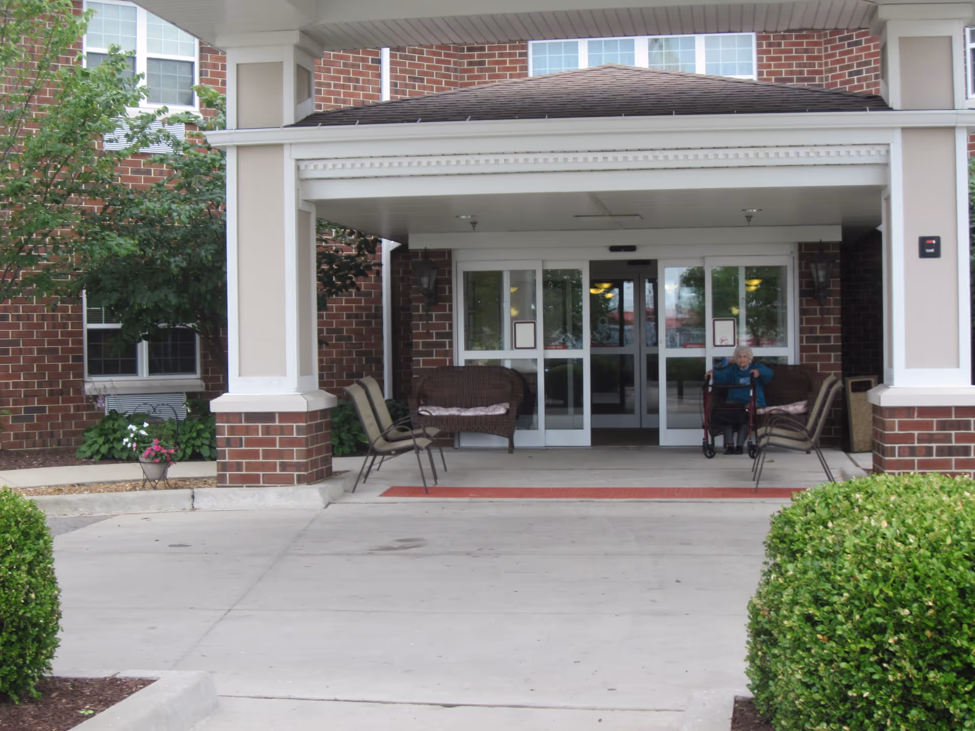 Entrance of a brick building with a covered drop-off area. There are chairs and a bench on either side of the entrance. An elderly person with a walker is sitting near the door. Green bushes and plants are visible around the entrance.