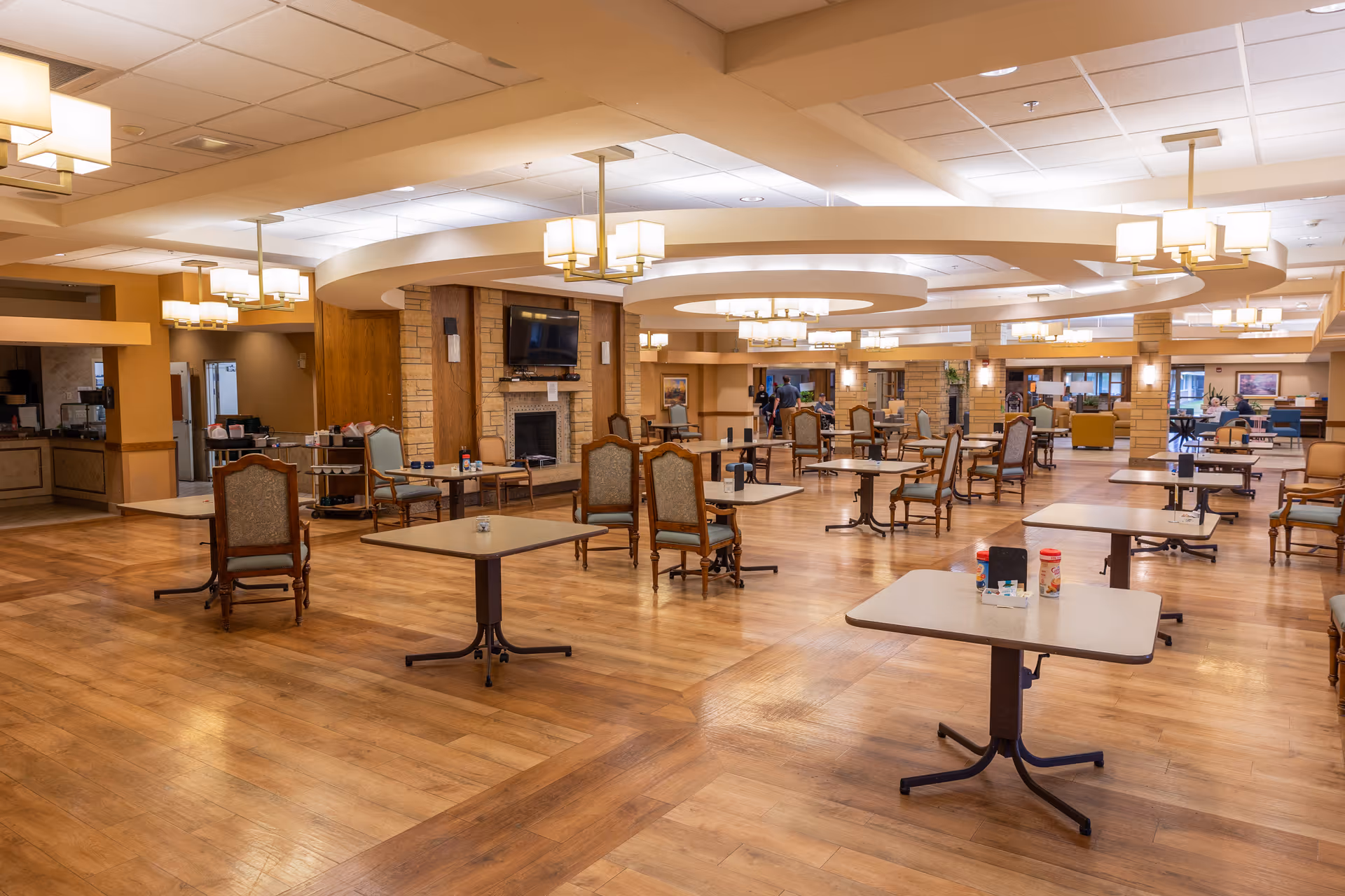 Spacious, well-lit communal dining and activity room with multiple tables and chairs, chandeliers, and a central fireplace with TV.