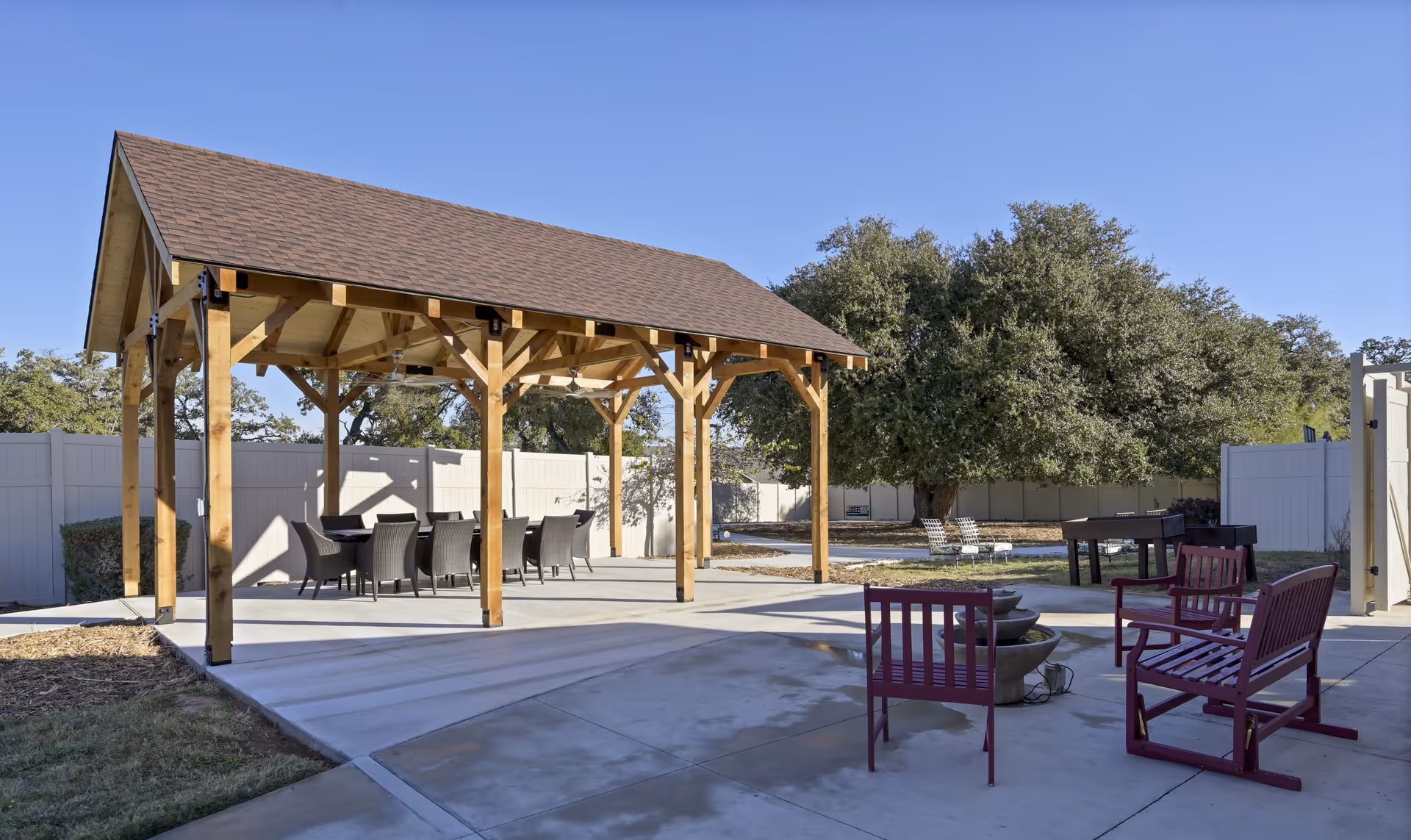 Outdoor patio area with a wooden pavilion covering a dining table and chairs, surrounded by a concrete patio with additional seating and a fire pit, large trees and a white fence in the background under a clear blue sky.