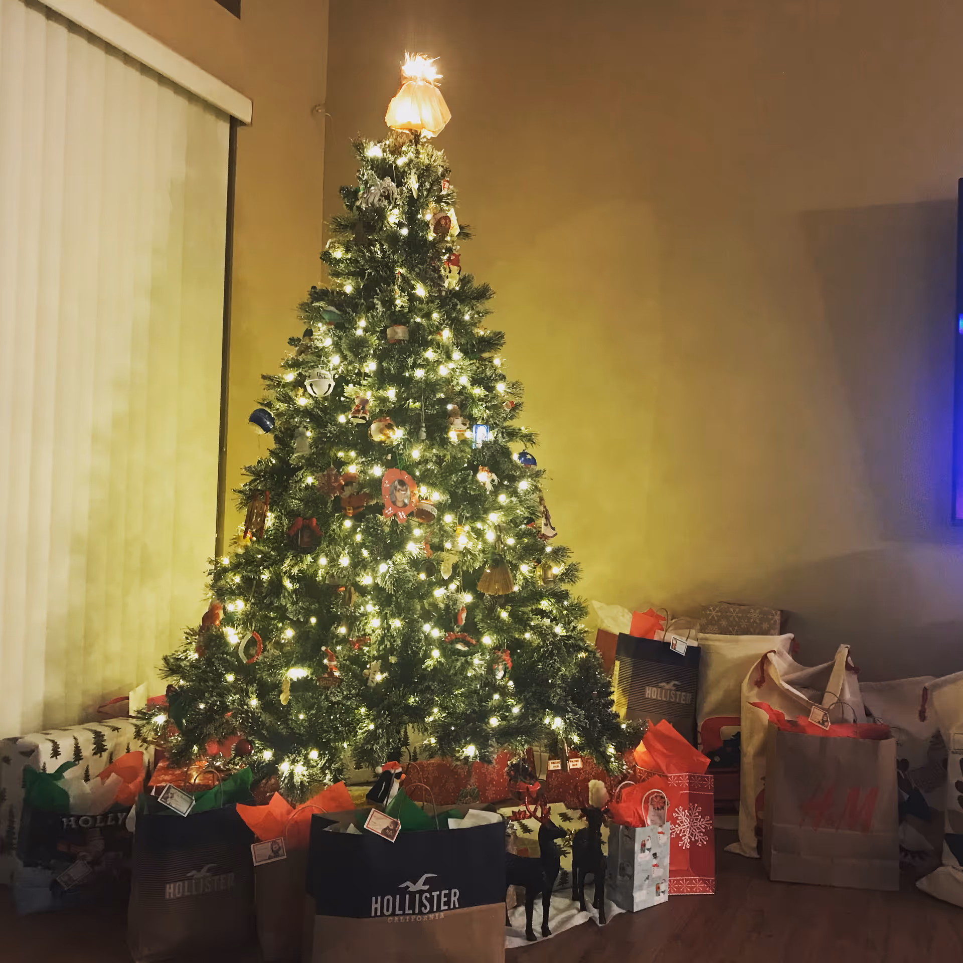 A decorated, lit Christmas tree in a living room surrounded by wrapped presents and gift bags.