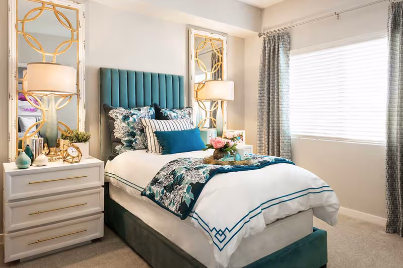 A neatly made bed with a teal upholstered headboard and floral bedding in a bright bedroom. Two decorative mirrors with gold accents hang above white nightstands on either side of the bed, each with a lamp and decorative items. A window with closed blinds and patterned curtains is on the right side of the room.